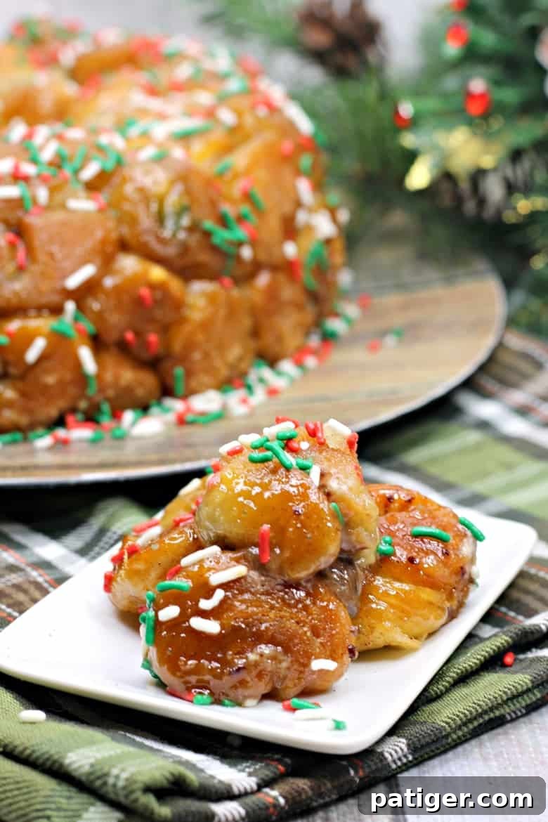 Yuletide Cinnamon Delight 2 Christmas morning monkey bread pieces coated in glaze and topped with red, green, and white sprinkles are arranged on a small white plate, placed in front of the full monkey bread ring on a wooden plate, ready to be pulled apart.