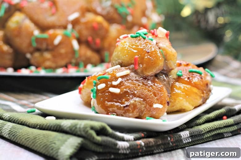 Yuletide Cinnamon Delight 9 Glossy Christmas monkey bread chunks, richly coated and topped with red, green, and white sprinkles, presented on a small white plate over festive green plaid fabric. A larger monkey bread ring is artfully blurred in the background.