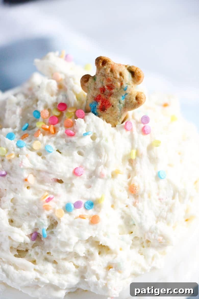 Creamy funfetti dip topped with pastel sprinkles with a teddy bear shaped cookie sticking out of it. A perfect dessert for parties.