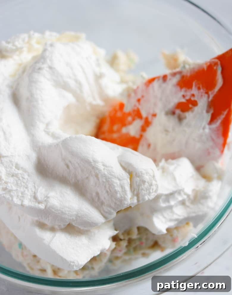 A large mixing bowl with a red spatula gently folding in the fluffy whipped topping into the vibrant cake batter dip mixture.