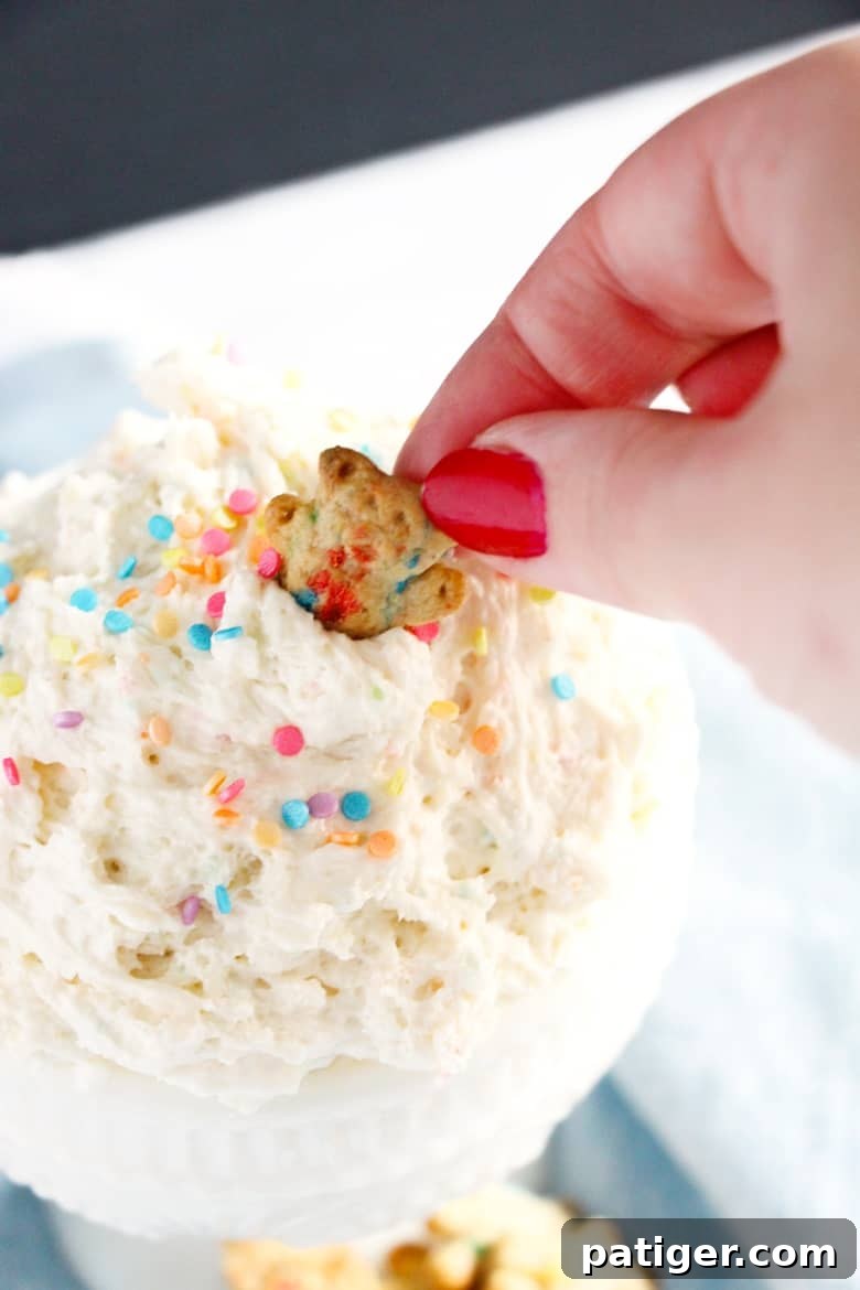 A hand dipping a cheerful teddy graham into a serving bowl of creamy funfetti cake batter dip, which is beautifully garnished with vibrant sprinkles.