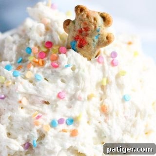 A teddy graham cookie being dipped into a luscious bowl of Funfetti cake batter dip, garnished with colorful sprinkles.