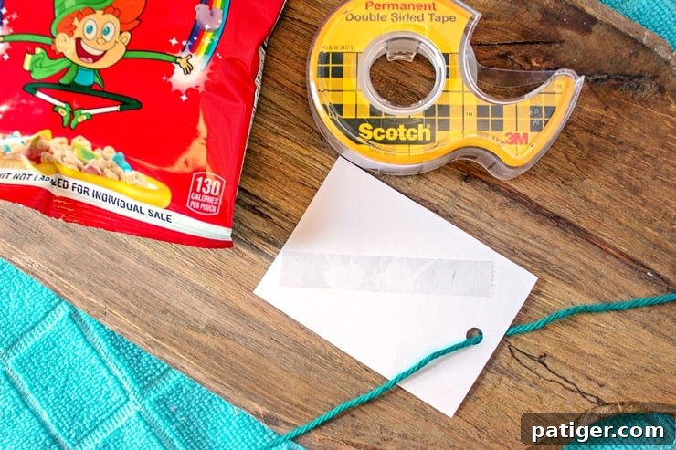 The back of a gift tag showing double-sided tape and threaded yarn, next to a roll of tape and a Lucky Charms bag