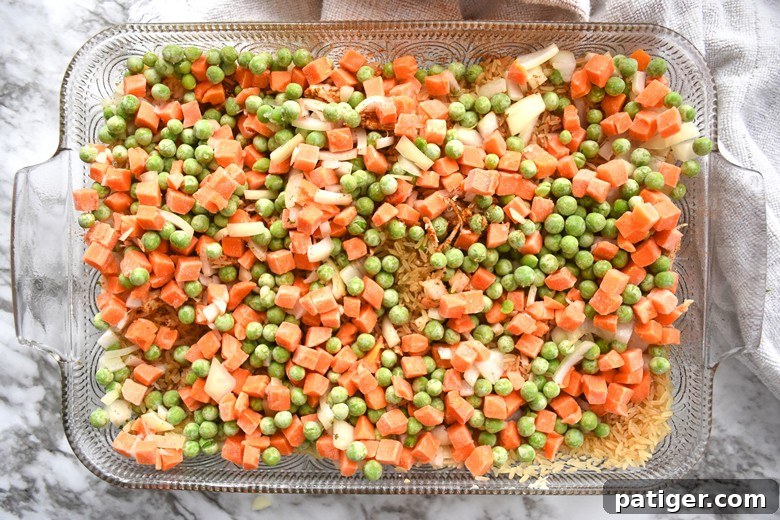 Layer of chicken topped with uncooked rice, frozen peas and carrots, and chopped onions in a baking dish