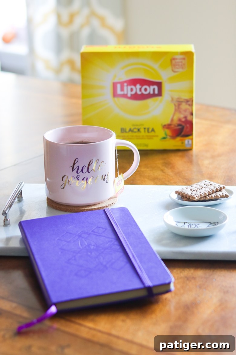Craft Your Own Tea Lovers Basket 7 A serene tea time setup featuring a 'Hello Gorgeous' pink coffee mug, a vibrant purple notebook, and delicate tea biscuits arranged on a tray, with a fresh box of Lipton tea in the background.