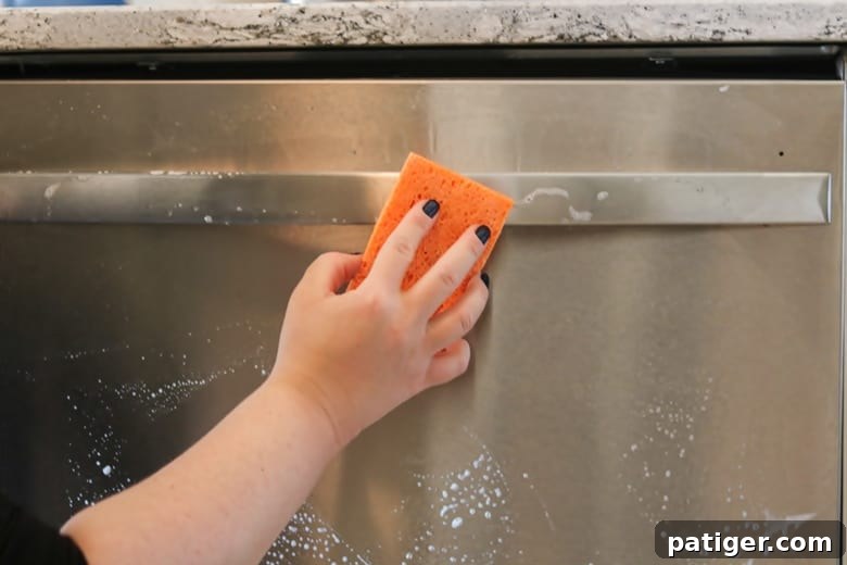 A hand holding a sponge and wiping a sudsy stainless steel dishwasher handle