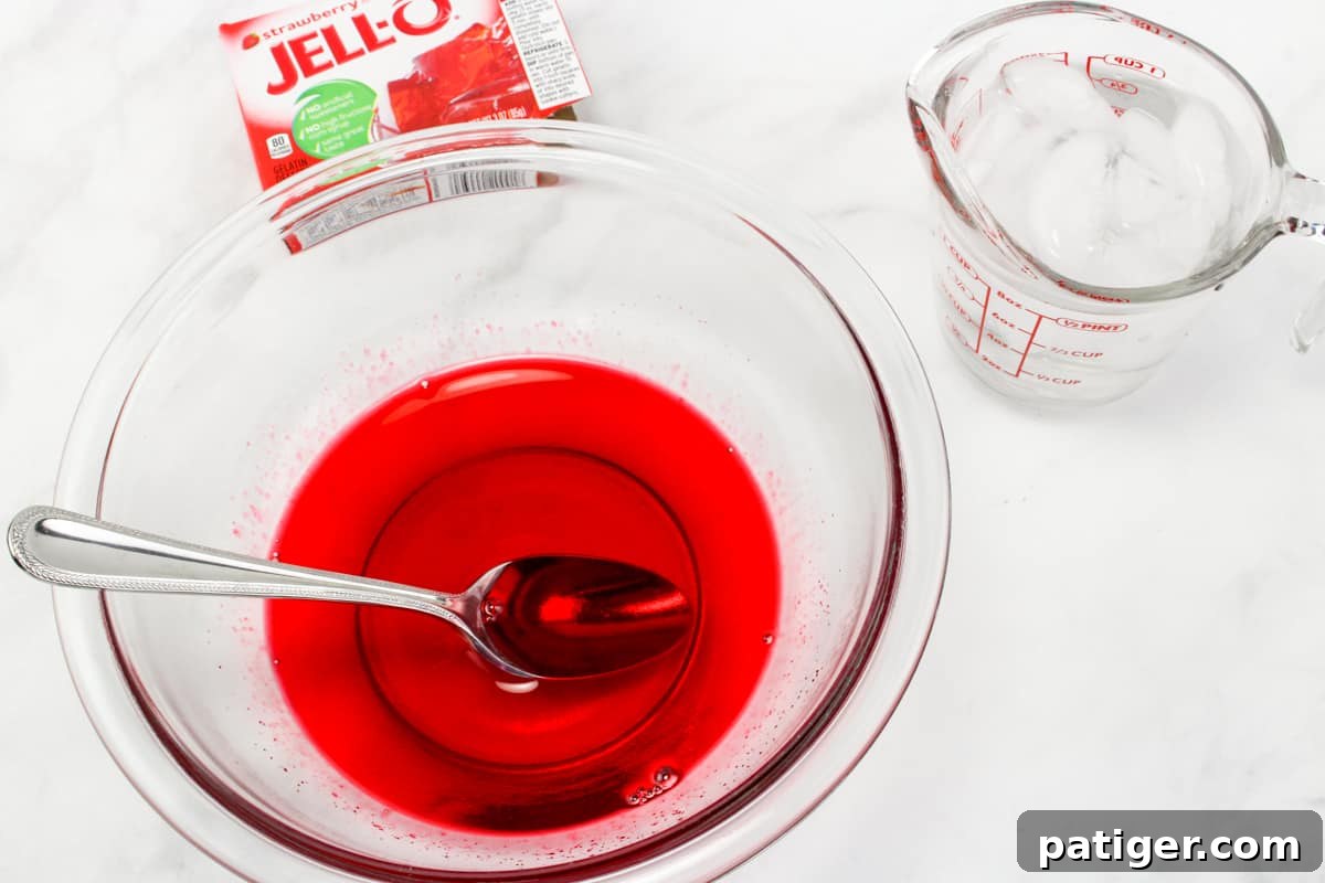 Bowl of jello mix dissolved in water with a spoon in it and box of strawberry jell in background.