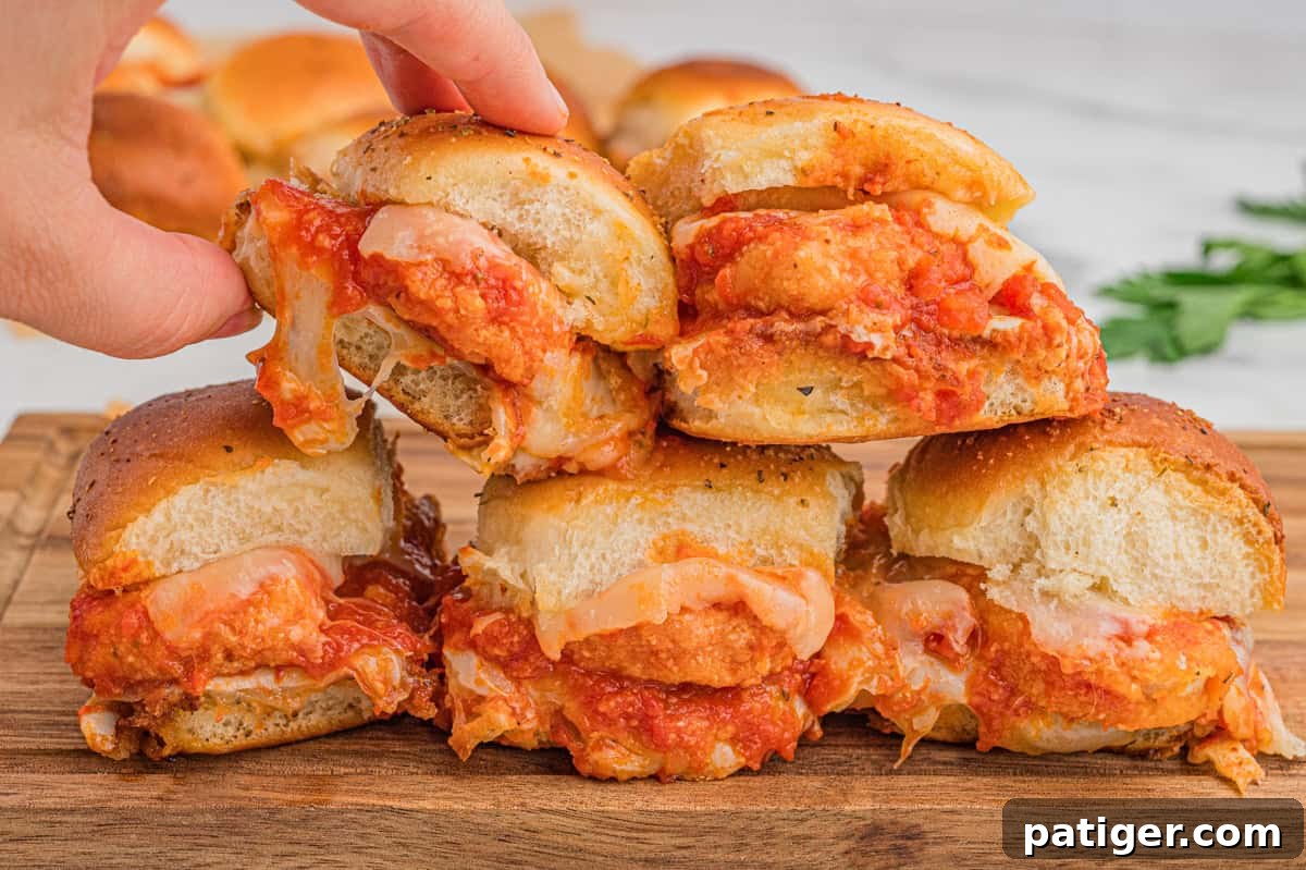 Chicken parmesan sliders are stacked on a wooden board. Each sandwich contains a breaded chicken nugget, marinara sauce, and melted cheese, with slightly toasted, herb-sprinkled buns. A hand is lifting one from the stack.