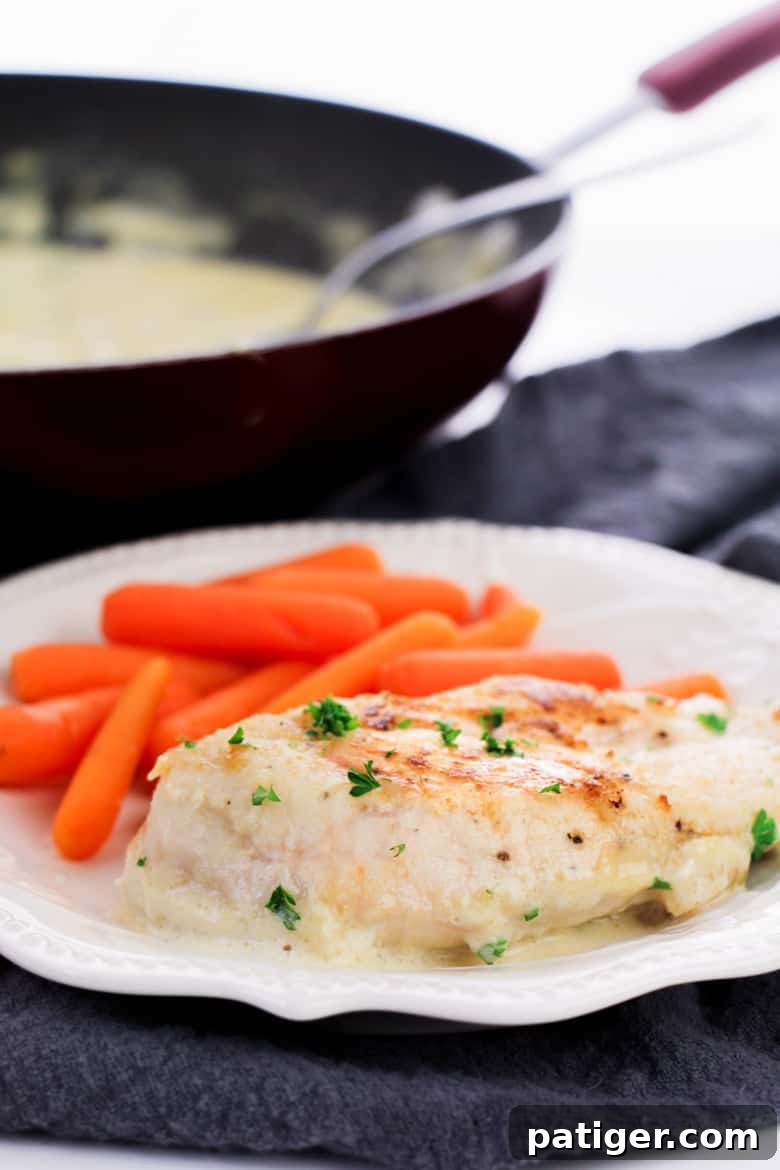 Creamy garlic chicken breasts on a white plate with baby carrots, skillet in background