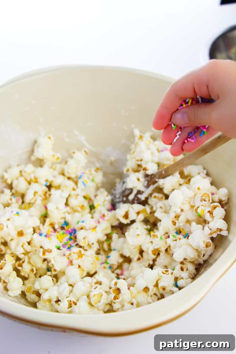 A hand gently adding a colorful cascade of rainbow sprinkles into a bowl of freshly coated popcorn, creating a funfetti effect for the cake batter treat.