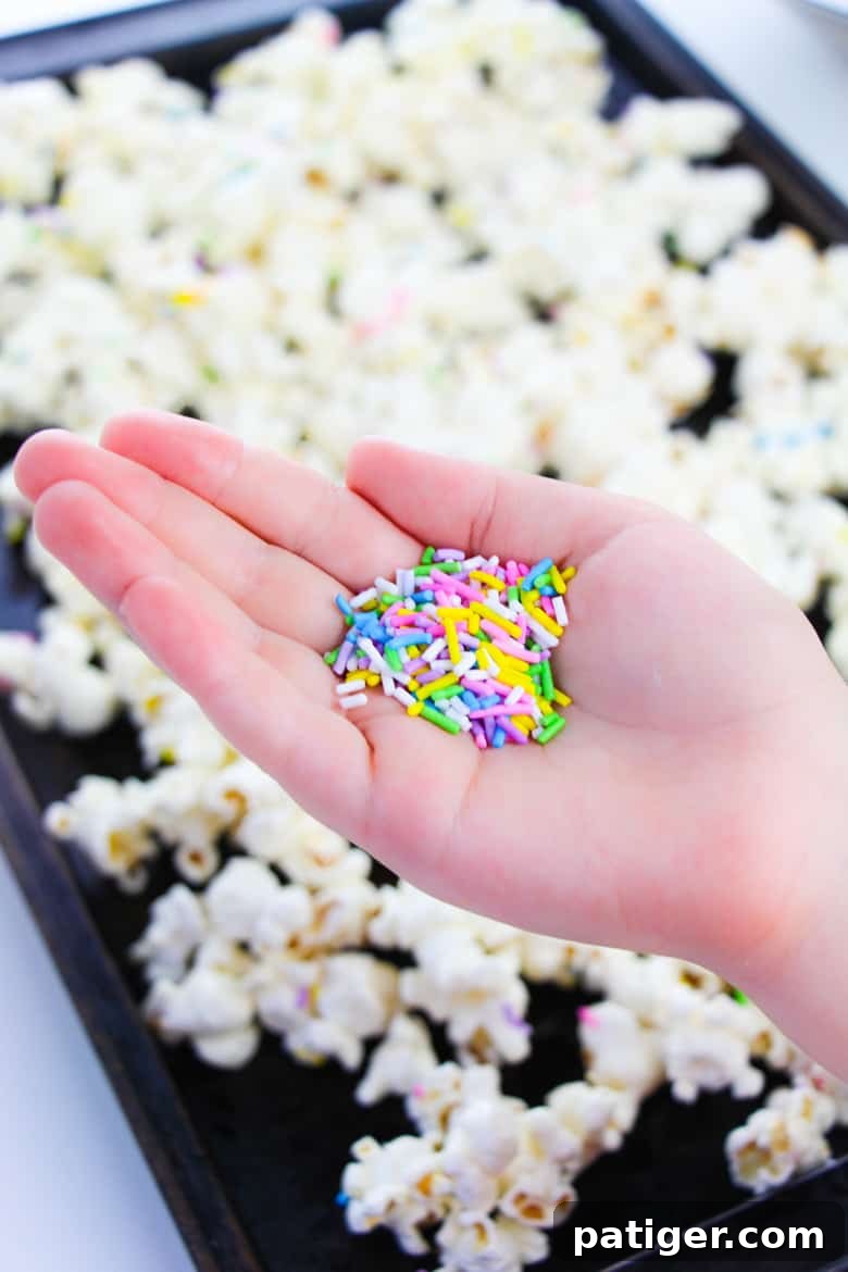 A woman's hand carefully holding a measuring spoon filled with vibrant rainbow sprinkles, poised to shower them over a tray of delightful cake batter popcorn.