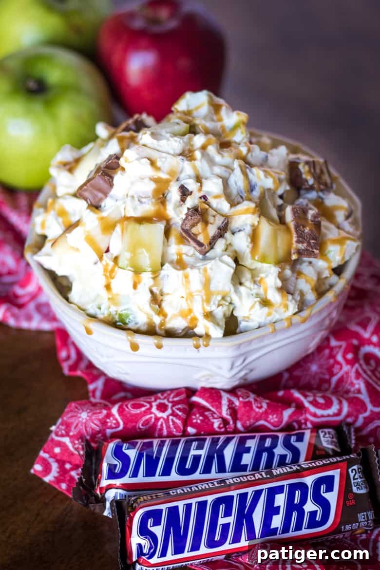 Ultimate Snickers Caramel Apple Salad 2 A beautifully presented Snickers Apple Salad in a pristine white bowl, with crisp apples and two Snickers bars visible in the background and foreground, setting the stage for a delightful dessert.