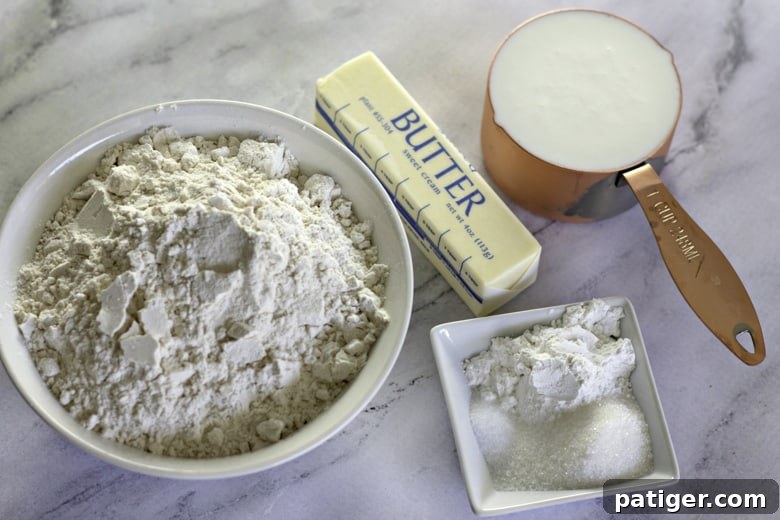 Fluffy Buttermilk Biscuits from Scratch 4 Ingredients for homemade buttermilk biscuits laid out on a counter: sticks of butter, a bowl of flour, baking powder, sugar, salt, and a glass of buttermilk.