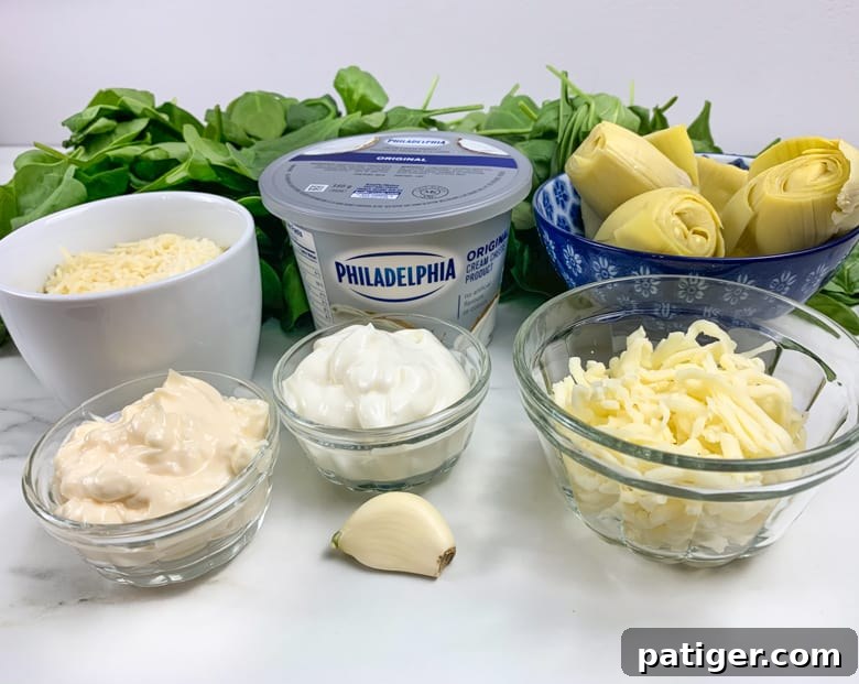 A flat lay photograph of all the ingredients for spinach artichoke dip: grated Parmesan, shredded mozzarella, cream cheese, sour cream, canned artichoke hearts, a generous pile of fresh spinach, and a single garlic clove.