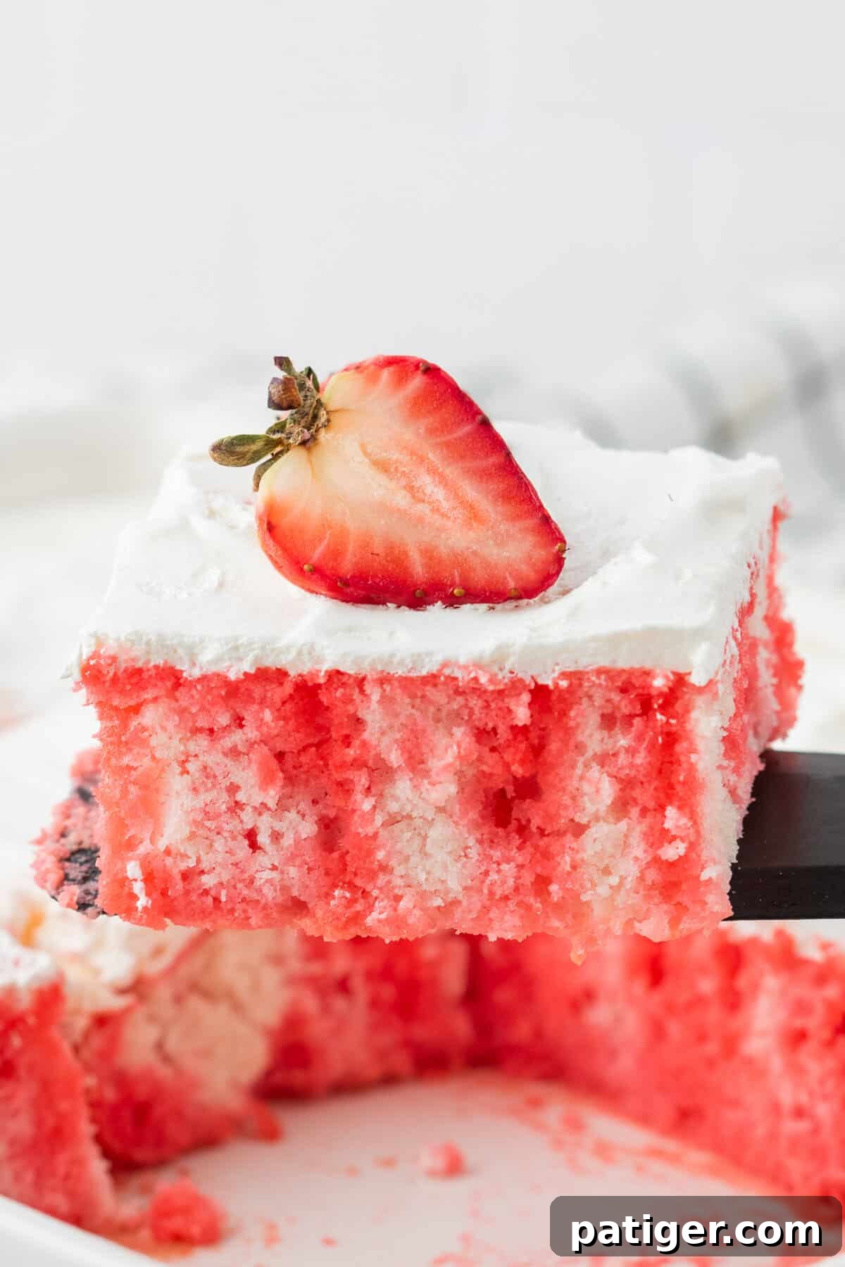 Strawberry Infusion Poke Cake 7 A spatula lifting a perfect slice of strawberry jello poke cake, showing the red Jello streaks, from a rectangular baking dish.