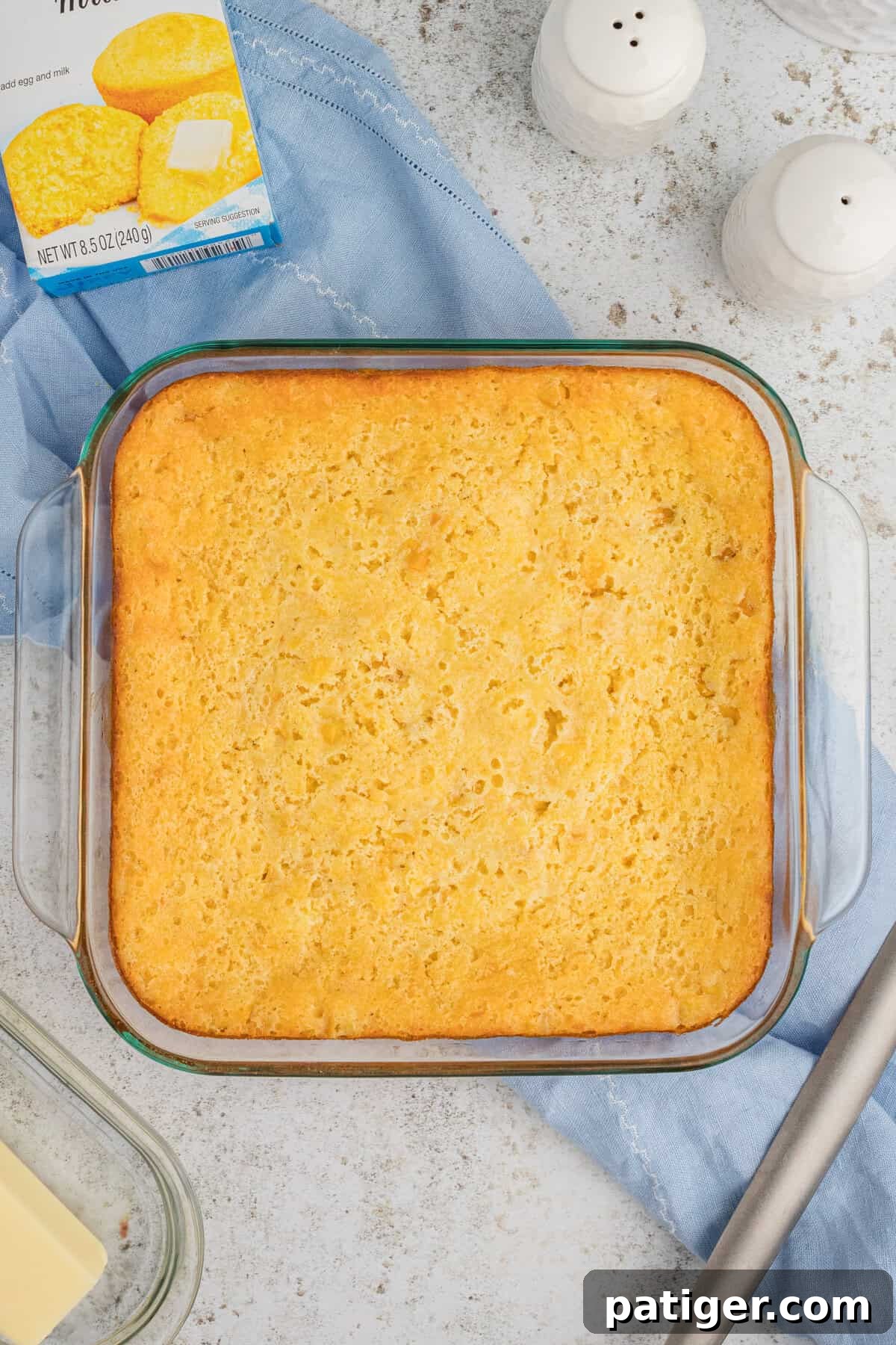 Golden Jiffy Cornbread and Creamed Corn Bake 3 A square glass baking dish filled with Jiffy Cornbread Casserole, with a box of Jiffy mix positioned beside it on a wooden surface.