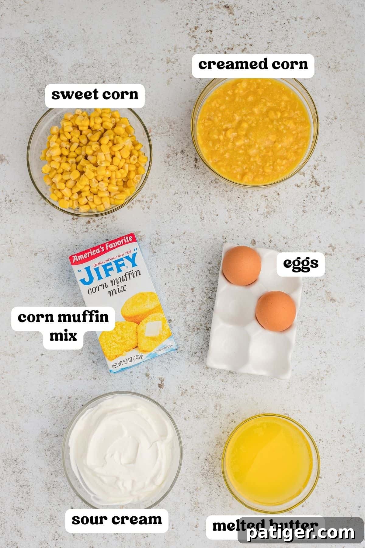 Golden Jiffy Cornbread and Creamed Corn Bake 4 Individual ingredients for Jiffy Cornbread Casserole laid out: a box of Jiffy mix, cans of creamed corn and whole kernel corn, two eggs, a bowl of sour cream, and melted butter.