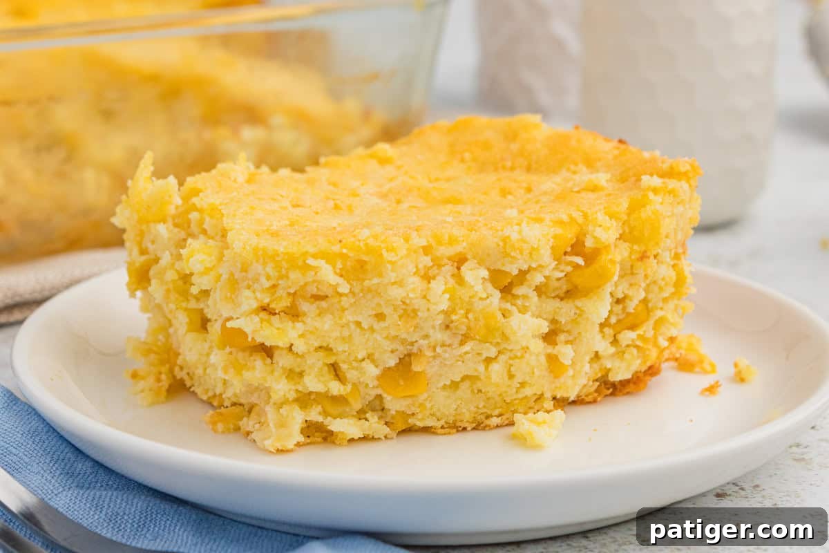 Golden Jiffy Cornbread and Creamed Corn Bake 7 A rectangular slice of Jiffy cornbread casserole, with visible corn kernels, is presented on a white plate.