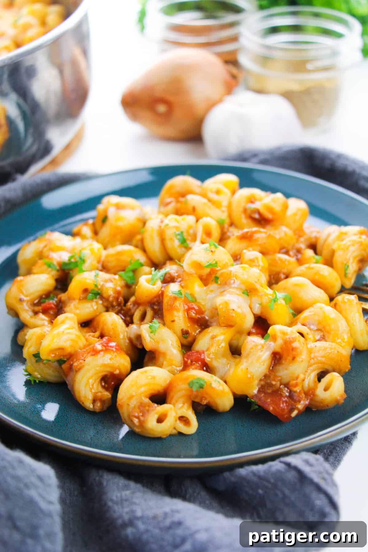 Blue plate with cheesy enchilada pasta topped with chopped cilantro. Onion, garlic, spice jars, and skillet full of pasta can be seen in background