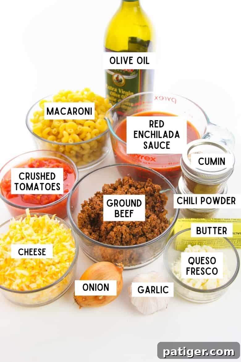 labeled recipe ingredients in glass bowls: macaroni, red enchilada sauce, cumin, chili powder, queso fresco, shredded cheese, crushed tomatoes, and ground beef. Also, a stick of butter, onion, garlic, and bottle of olive oil
