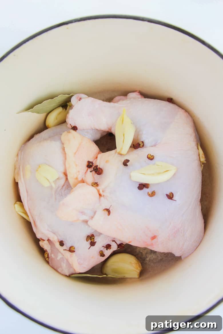 Raw chicken quarters nestled in a large Dutch oven, surrounded by crushed garlic cloves, whole peppercorns, and various seasonings, ready for poaching.