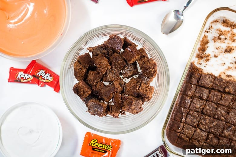 A close-up image showing hands assembling a Halloween Candy Trifle, layering brownie pieces, orange pudding, and whipped topping in a clear trifle dish.
