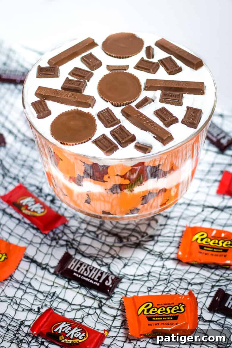A close-up, top-down view of a completed Halloween Candy Trifle, showcasing rich chocolate brownies, vibrant orange pudding, and white whipped topping, garnished with various HERSHEY® Halloween candies.