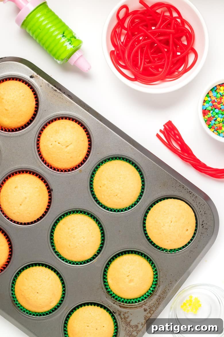 Cooled vanilla cupcakes in a pan next to green frosting in a piping bag, red string candy, and sprinkles