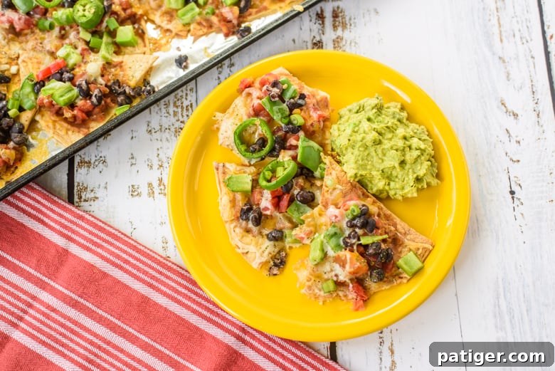Platter of vegetarian nachos with beans, peppers, and melted cheese, served with guacamole on the side. More nachos visible on a sheet pan in the background.