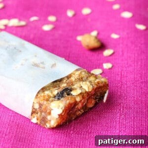Homemade granola bar wrapped in parchment paper with end exposed. Rolled oats are sprinkled on the pink tablecloth in the background.