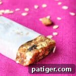 Homemade granola bar wrapped in parchment paper with end exposed. Rolled oats are sprinkled on the pink tablecloth in the background.