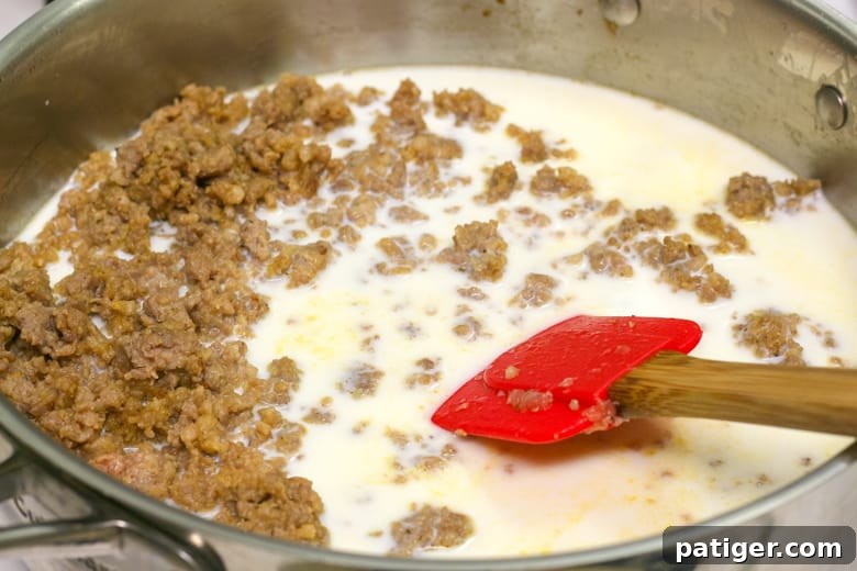 A skillet filled with cooked ground sausage, with milk being slowly poured in and a spatula for stirring.