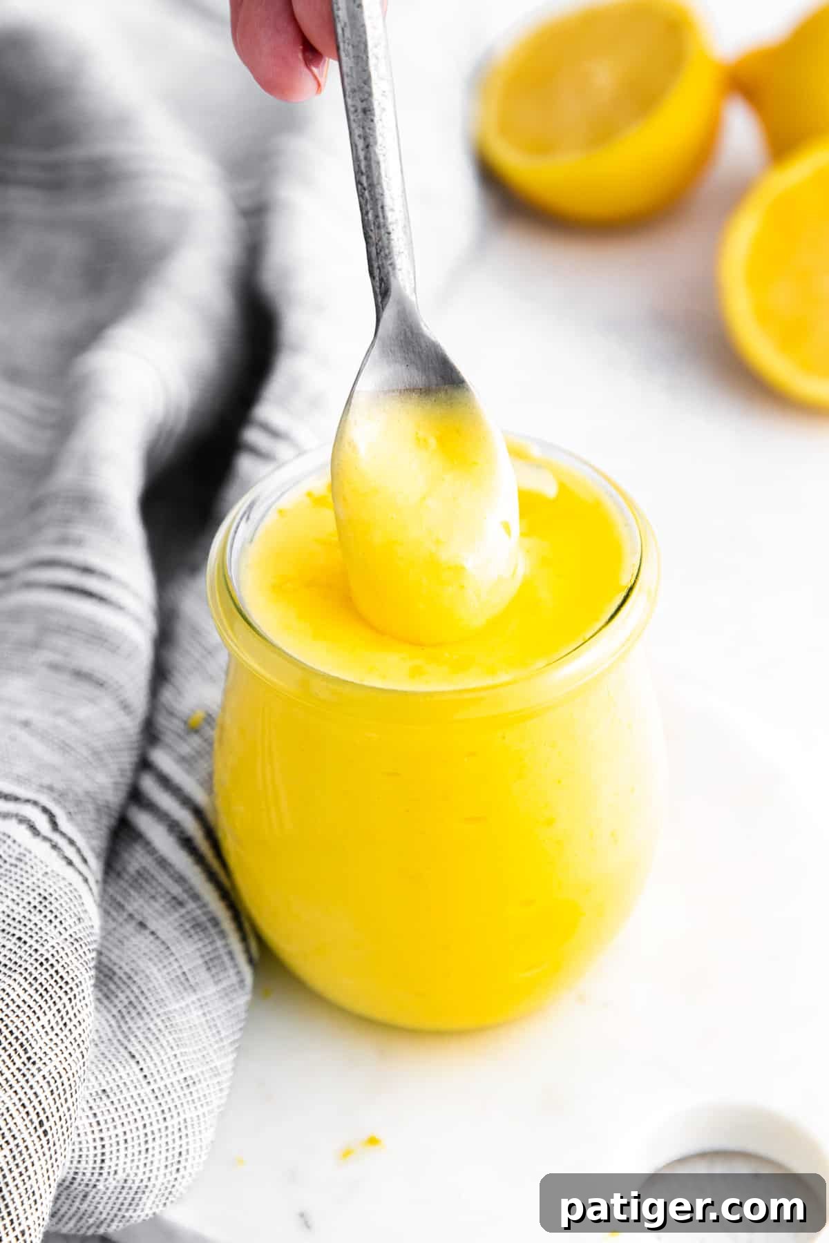 Small glass jar of bright yellow lemon curd; a silver spoon is pulling out a spoonful from the top to show the creamy texture. Lemon halves and grey linen in background.