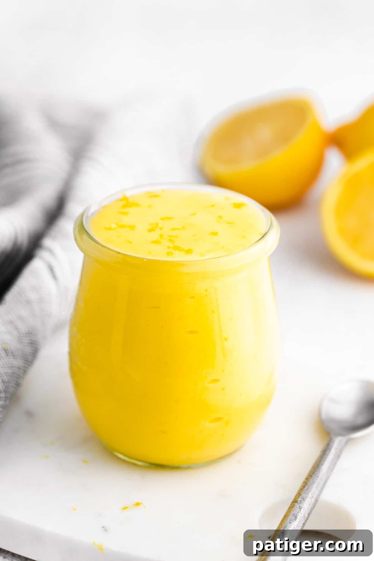 Lemon curd in a glass jar, with lemon halves and a silver spoon next to it