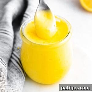 Small glass jar of bright yellow lemon curd; a silver spoon is pulling out a spoonful from the top to show the creamy texture. Lemon halves and grey linen in background.