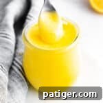 Small glass jar of bright yellow lemon curd; a silver spoon is pulling out a spoonful from the top to show the creamy texture. Lemon halves and grey linen in background.