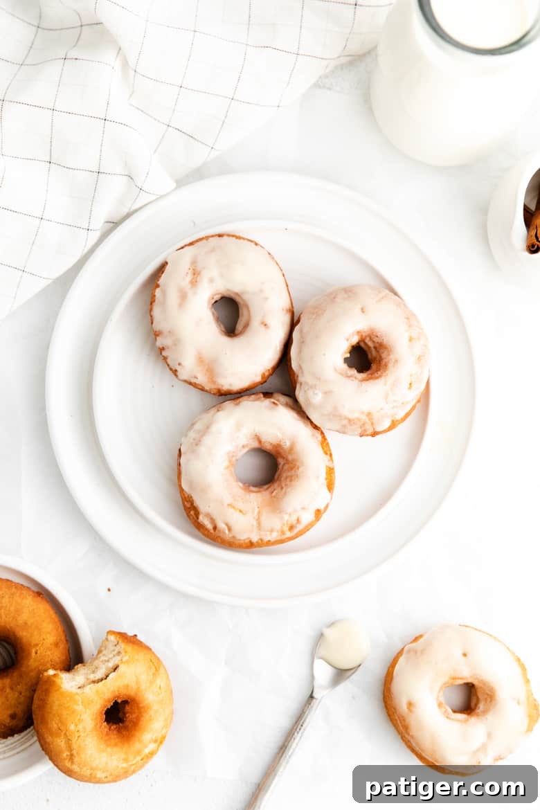 Three glazed cake doughnuts on a white plate surrounded by a white linen towel, glass jug of milk, a spoon, and additional doughnuts, some un-glazed