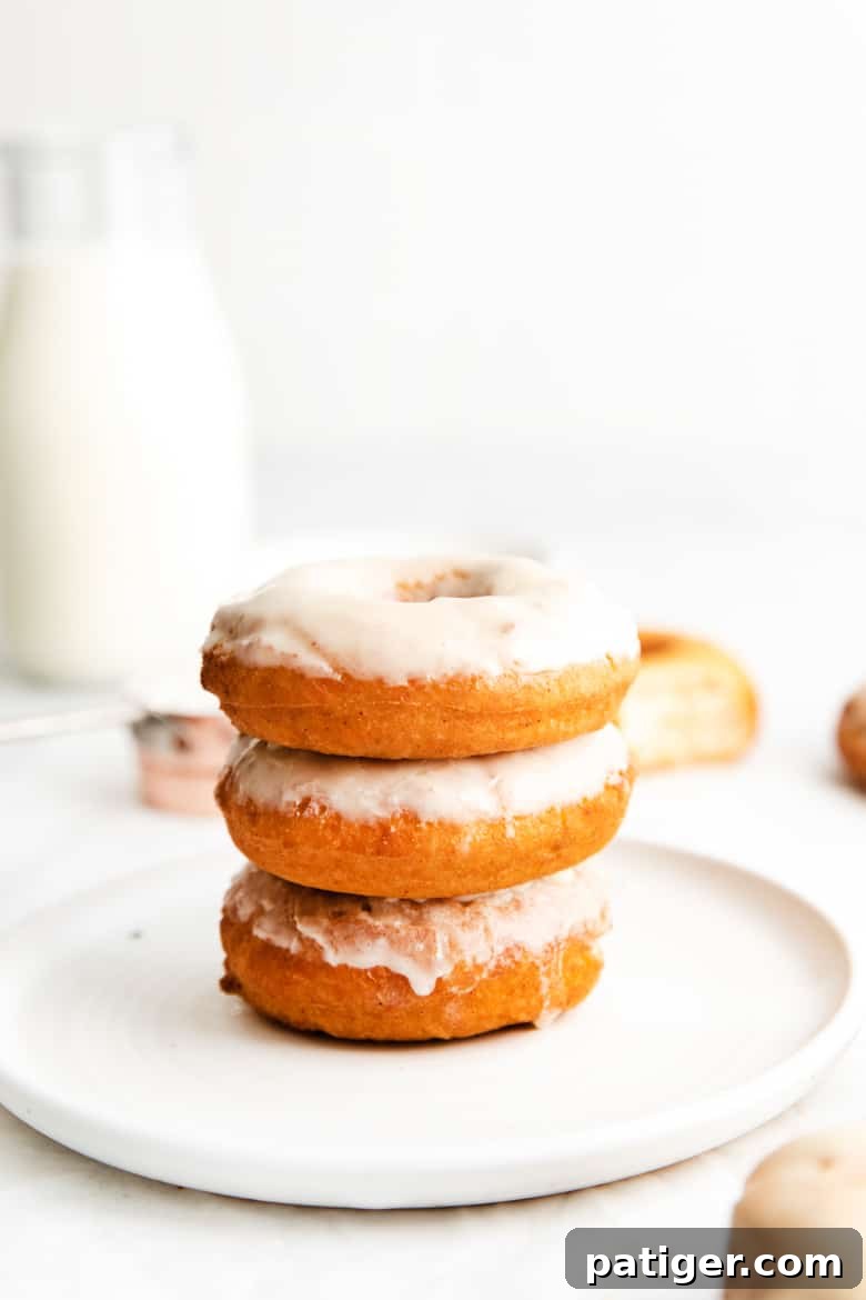 Three glazed doughnuts stacked on top of one another, on a white plate. A glass milk jug filled with milk is in background