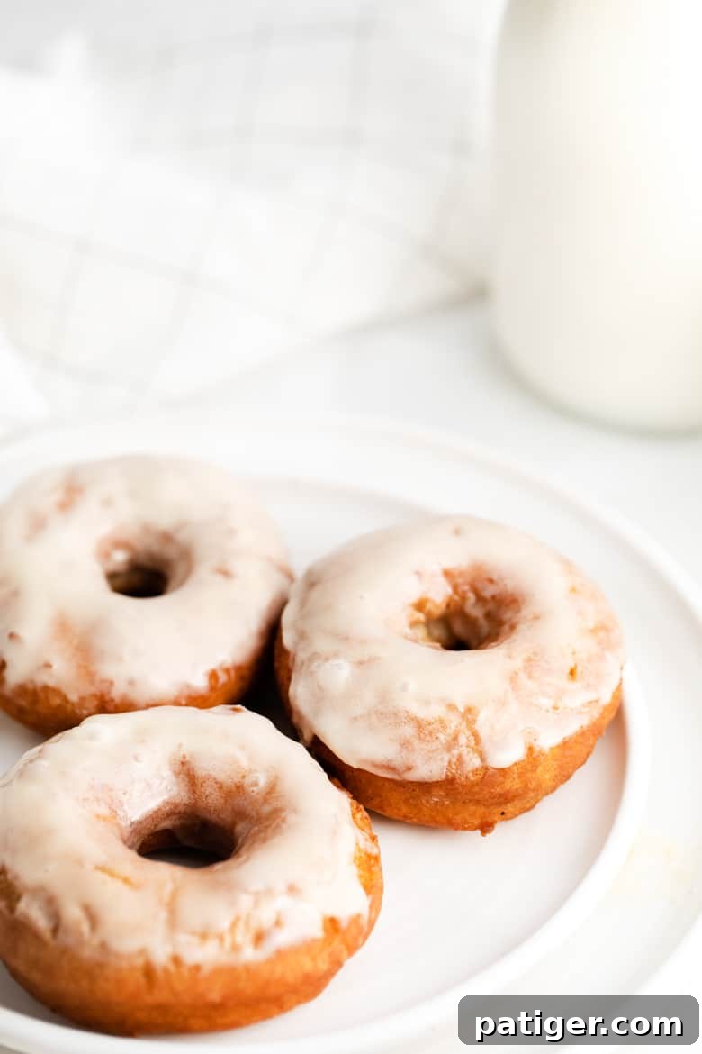 Three glazed donuts on a white plate with a glass container of milk next to them