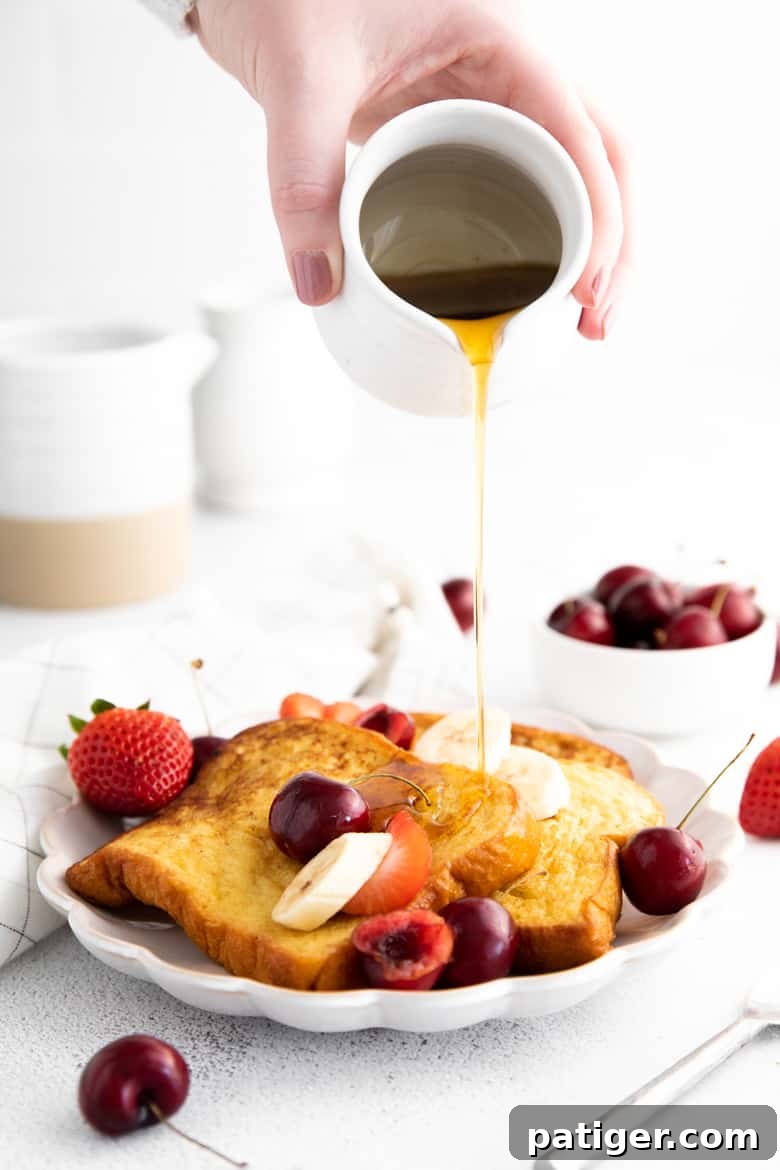 Hand pouring pitcher of maple syrup over french toast with topped with fresh fruit. Bowl of cherries in background