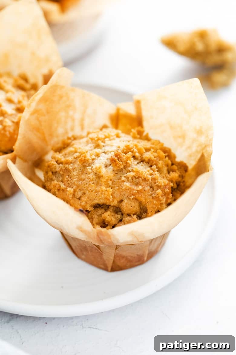 Crumb cake muffin in brown wrapper on a white plate, showcasing its golden crumb topping and soft interior.