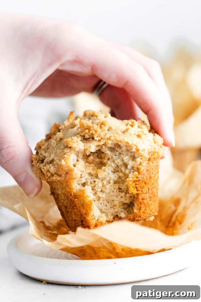 Hand holding a coffee cake muffin with a bite taken out, revealing the soft, cinnamon-vanilla interior and the delicious streusel topping.