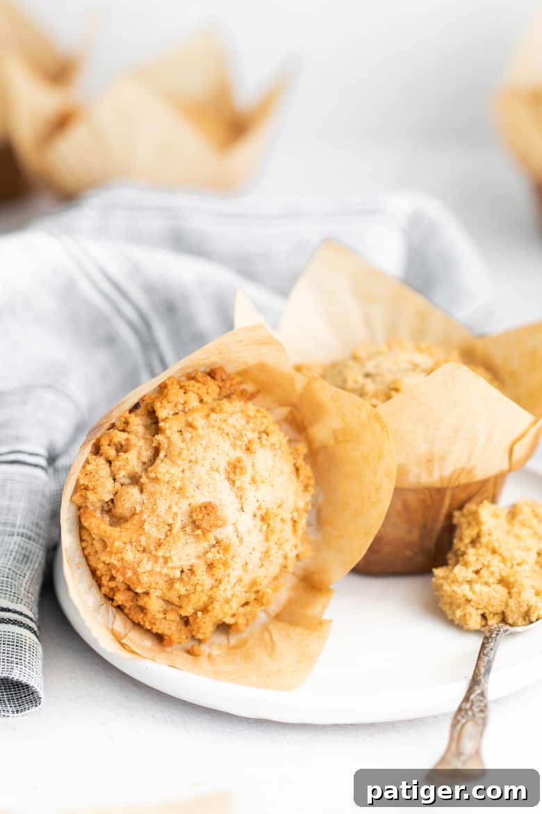 A wrapped crumb cake muffin, a spoonful of crumb topping, and an unwrapped muffin sit on a white plate, highlighting the delightful streusel.