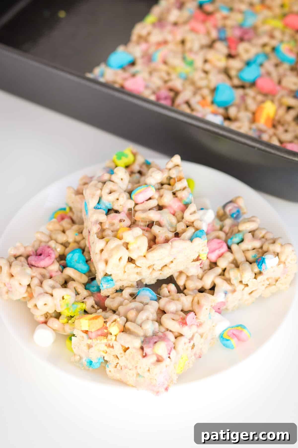 Magical Lucky Charms Treats 3 Lucky charms bars cut into squares and piled on a white plate. Baking dish with uncut treats is behind them.