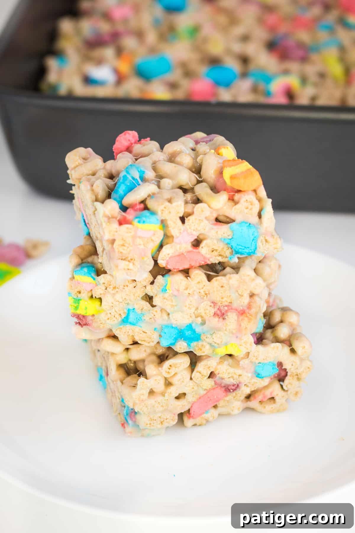 Magical Lucky Charms Treats 8 Three lucky charms marshmallow cereal bars cut into squares and stacked on top of one another on a white plate. A baking pan with uncut treats is behind them.