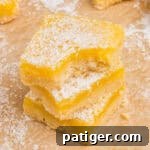 3 square lemon bars with shortbread crust and dusting of powdered sugar stacked. The top bar has a bite taken out of it.