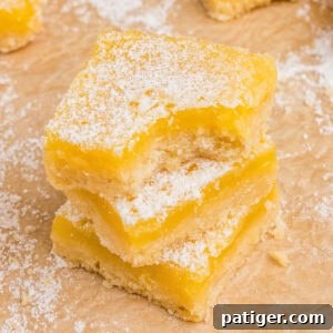3 square lemon bars with shortbread crust and dusting of powdered sugar stacked. The top bar has a bite taken out of it.