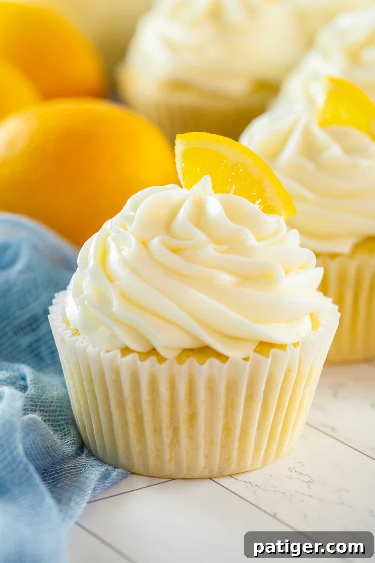 Lemon cupcakes with cream cheese frosting and fresh lemon slices for garnish, artfully arranged with whole lemons and a blue napkin in the background.
