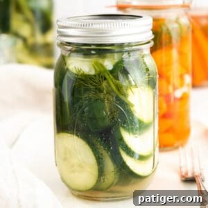 Wide Mouth Mason Jar filled with sliced pickled cucumbers. More jars in background, some with pickled cucumbers and others with carrots.