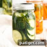 Wide Mouth Mason Jar filled with sliced pickled cucumbers. More jars in background, some with pickled cucumbers and others with carrots.
